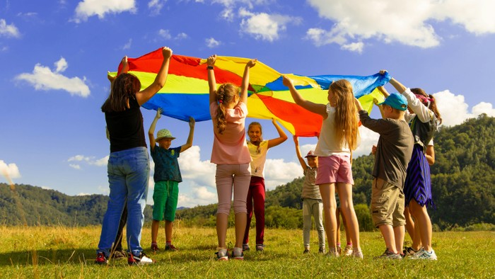 Eine Gruppe von Kindern und Erwachsenen hält fröhlich ein großes, buntes Fallschirmtuch auf einem offenen Feld.