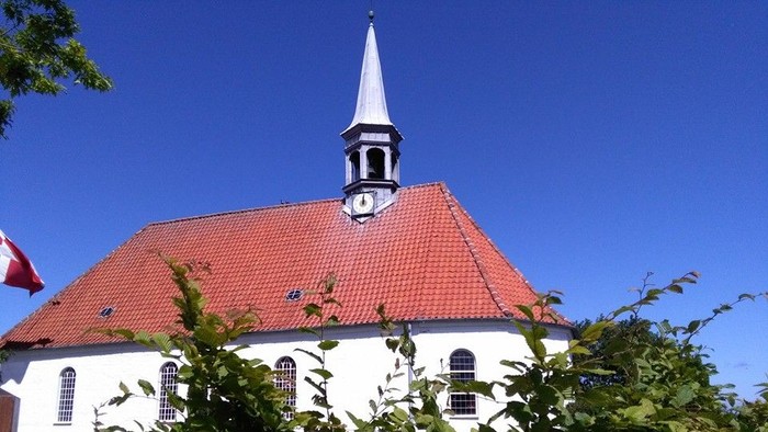 En charmerende hvid kirke med rødt tag og spir omgivet af frodigt grønt løv under en klar blå himmel.