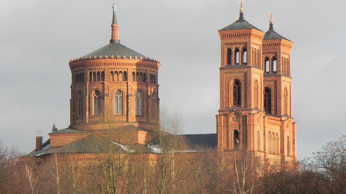 Zwei Türme einer Kirche mit spitzen Dächern und goldenen Akzenten, umgeben von Bäumen und einem bewölkten Himmel