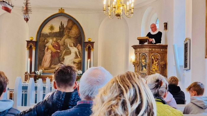 Et menighedshold lytter opmærksomt under en gudstjeneste, hvor en præst læser op fra en prædikestol.