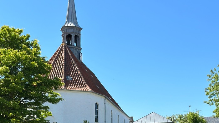 Hyggelig hvid kirke med frodig have og højt spir under klar blå himmel.
