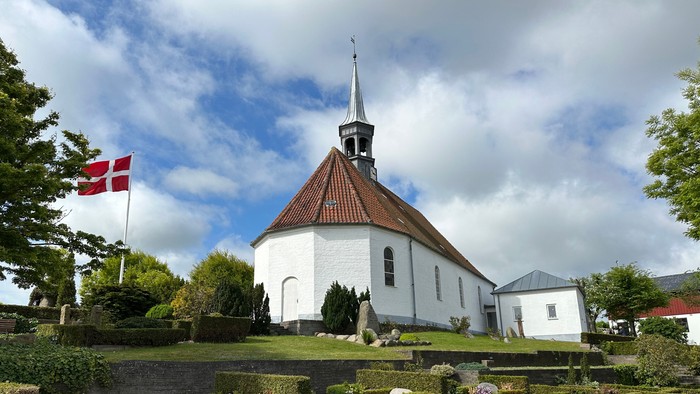 En hvid kirke med tårn og en dansk flag på en bakke med gravsteder og buske.