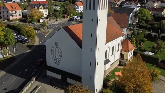 Weiße Kirche mit hohem Glockenturm und Kreuz, umgeben von Einfamilienhäusern und Bäumen.