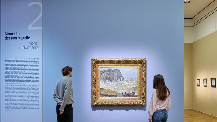 Zwei Personen betrachten Monets Landschaftsgemälde der Normandie in einer Kunstausstellung mit einer Informationsanzeige neben sich.