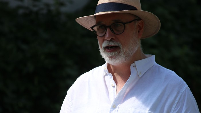 Ein Mann mit Hut und Brille hält ein Buch im Freien und wirkt nachdenklich und entspannt.