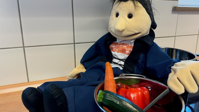 Ein Puppenkoch sitzt neben einem Topf voller Gemüse und bereitet eine Mahlzeit zu.