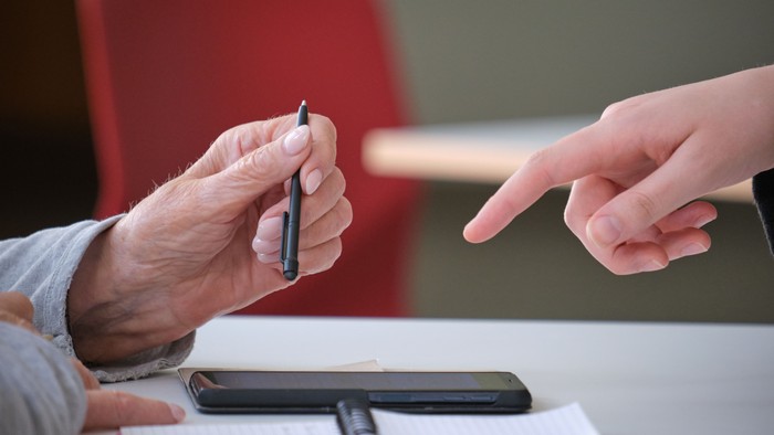 Zwei Personen tauschen einen Stift über einem Tisch mit Unterlagen und einem Smartphone aus.
