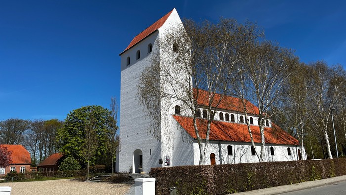 Historisk hvid kirke med rødt tegltag og et højt firkantet tårn omgivet af træer.