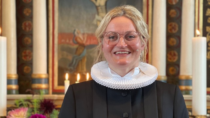 En person i formelt kirkeligt tøj står og smiler i en kirke, pyntet med blomster og religiøs kunst.