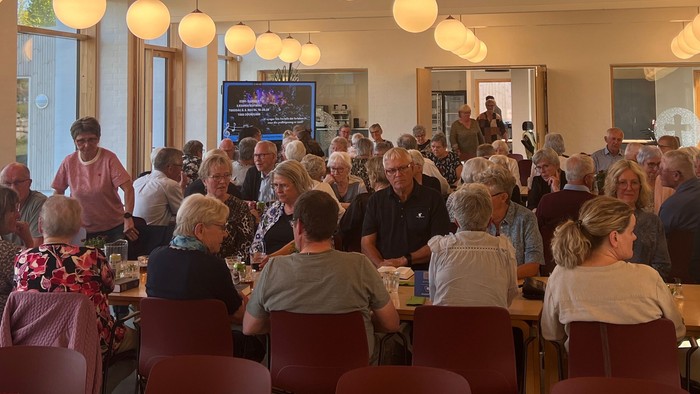 En livlig forsamling af mennesker siddende ved borde i en veloplyst, rummelig spisesal med moderne belysning.