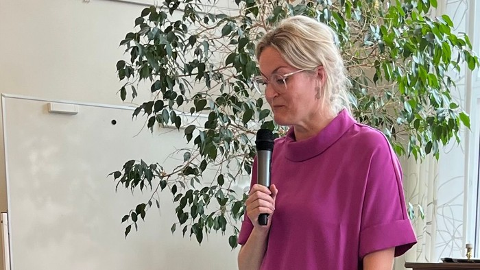 En person taler ind i en mikrofon ved et lille podium i et lokale med musikinstrumenter.