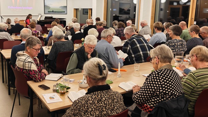 En gruppe ældre mennesker samledes i et lokale til et møde eller en diskussion med borde og præsentationer.