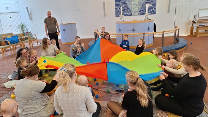 Gruppe af børn og voksne, der sidder i en cirkel og leger med store farverige faldskærme indendørs.