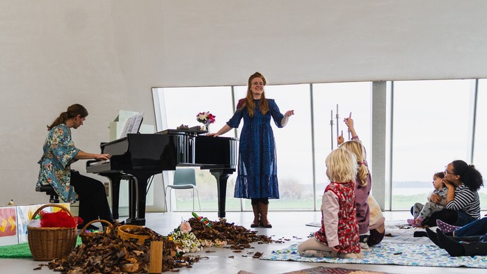 Kreativ workshop med musikere, der spiller klaver, børn, der laver håndværk, og spredte naturlige materialer på gulvet.