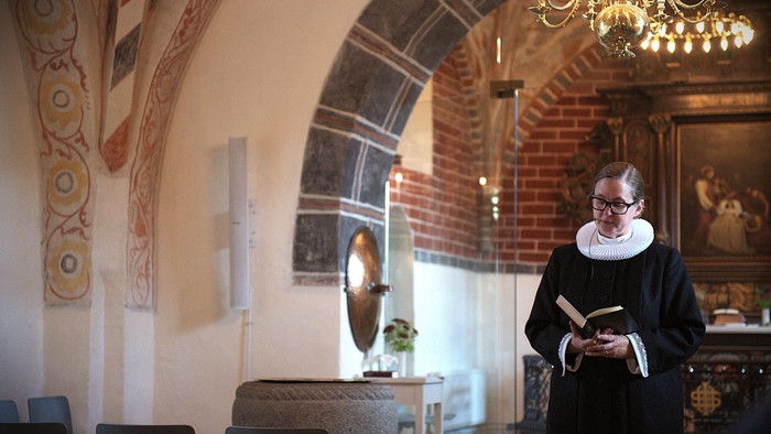 En nonne læser en bog i et historisk, rigt udsmykket kirkeinteriør med hvælvede murstensværker.