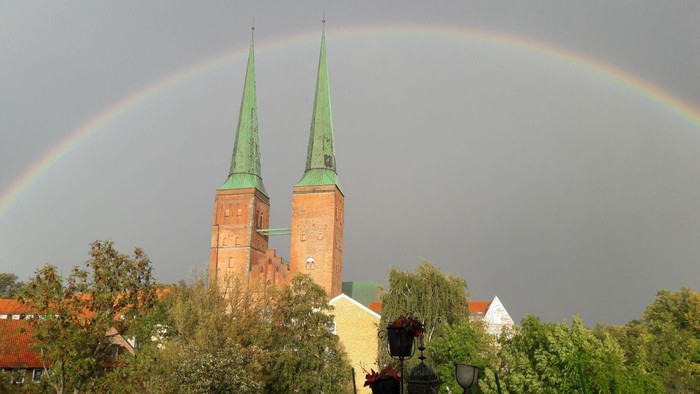 Ein lebendiger Regenbogen wölbt sich über zwei grünbedachte Kirchtürme, eingerahmt von üppigem Grün und einem bewölkten Himmel.