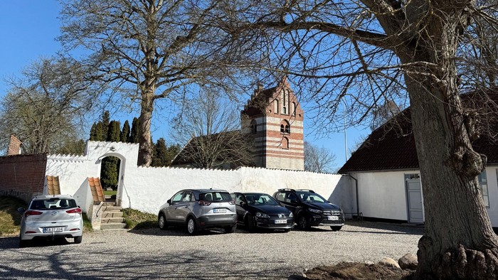 En fredelig gårdsplads med parkerede biler, en hvid mur og et højt træ under en klar blå himmel.