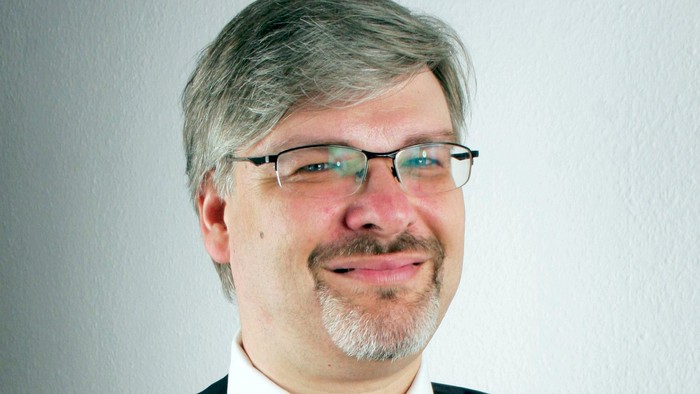 Professioneller Mann mit Brille, Weste, weißem Hemd und roter Krawatte, der leicht lächelt.