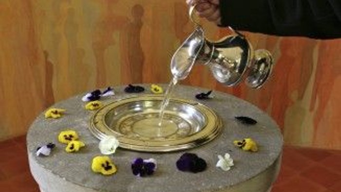 Eine Hand gießt Flüssigkeit in eine Schale auf einem steinernen Tisch, umgeben von bunten Blumen.
