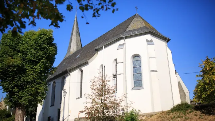 Eine malerische weiße Kirche mit einem steilen Turm, die vor einem klaren blauen Himmel steht und von Grün umgeben ist.
