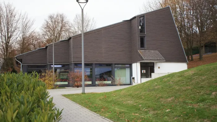 Modernes Gebäude mit schrägem Dach und großen Fenstern, das auf einem grasbewachsenen, gestalteten Hang liegt.