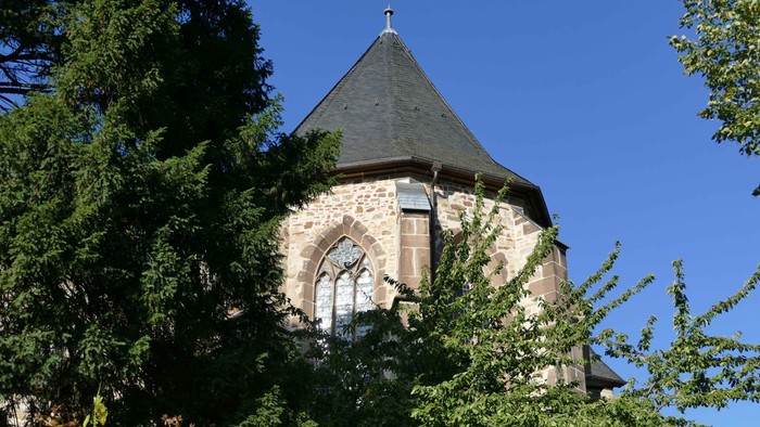 Steingebäude mit Rundbogenfenstern und spitzem Dach, teilweise von Bäumen verdeckt.