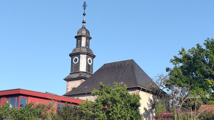 Historische Kirche mit hohem Kirchturm und Uhr, umgeben von Grünflächen und angrenzenden Gebäuden.