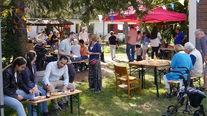 Im Freien sitzen Menschen an Tischen, einige stehen, es gibt Sonnenschirme und eine Kinderwagen.