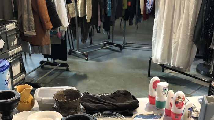 Ein überfüllter Marktstand mit Kleidung, Küchenutensilien und Putzmitteln auf Tischen und Ständern.
