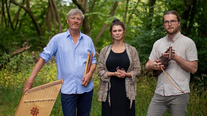 Tre musikere, der holder traditionelle strengeinstrumenter i en frodig, skovagtig udendørs omgivelse.