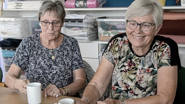 To ældre kvinder sidder ved bordet med tæpper og tøj i baggrunden