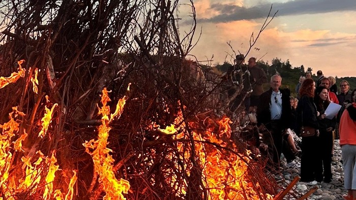 En gruppe mennesker står omkring en stor bål på en stenstrand ved solnedgang
