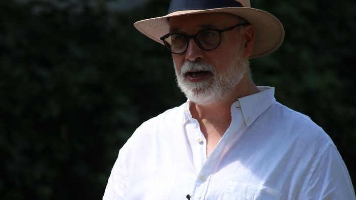 Ein Mann mit Hut und Brille hält ein Buch im Freien und wirkt nachdenklich und entspannt.