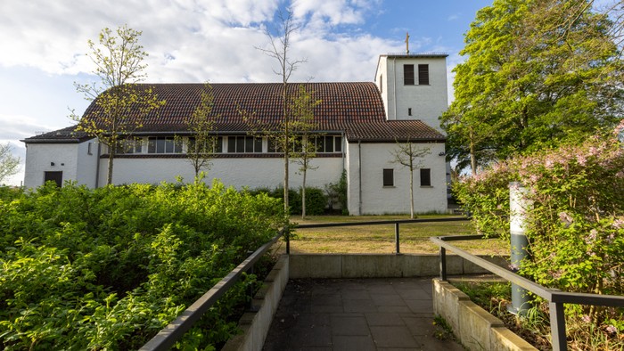 Einfache Kirche, nach dem Krieg erbaut, mit weißen Wänden, rotem Ziegeldach und üppigem Grün, das einen gepflasterten Weg umgibt.