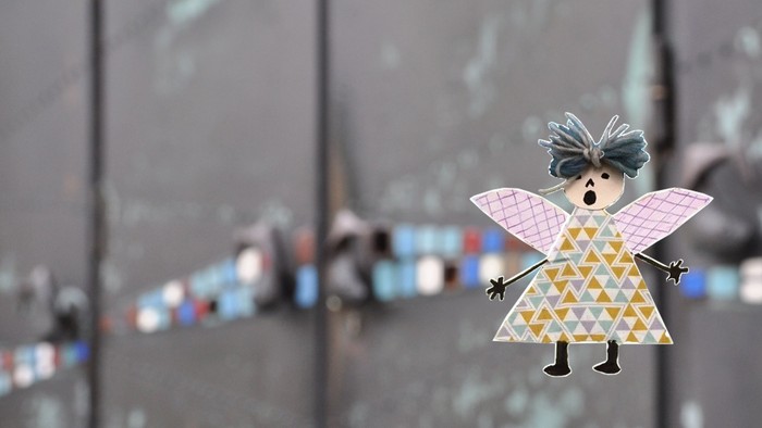 Bunte Papierengel-Dekoration mit weit geöffnetem Mund und ausgebreiteten Flügeln auf einer reflektierenden Oberfläche.