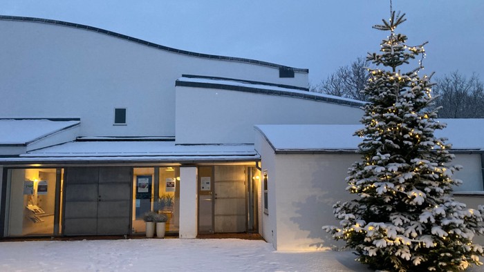 En snedækket juletree foran et belyset hus med flade tag.