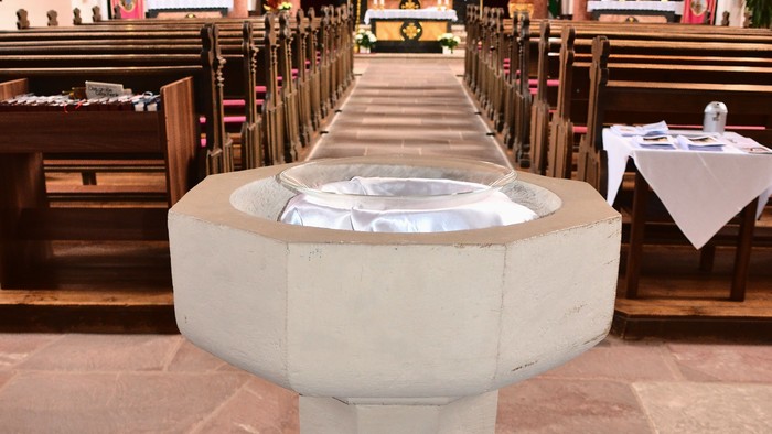 Steinerner Taufstein in einem Kirchenschiff, das zum Altar führt, mit hölzernen Kirchenbänken.