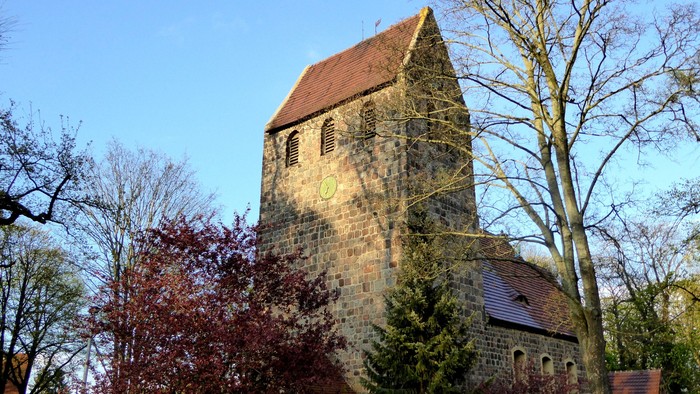 Alte Kirche mit hohem Turm und rotem Dach, umgeben von Bäumen und Büschen
