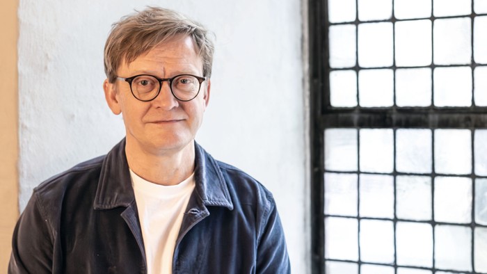 En mand med briller og en denimjakke står nær et vindue med et metalgitter.