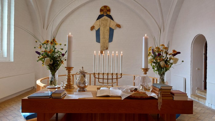 Elegant kirkealter med lys, blomster, religiøse bøger og et roligt korsfæstelsesmaleri ovenover.