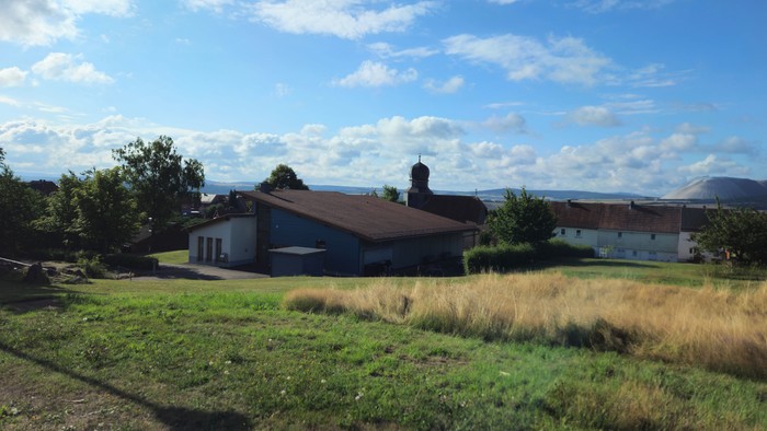 Ländliche Szene mit einem Haus und offenen Feldern unter einem hellen, teilweise bewölkten Himmel.