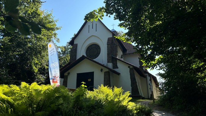 Kleine weiße Kirche mit Rundbogenfenster und Kreuz, umgeben von Bäumen und Büschen