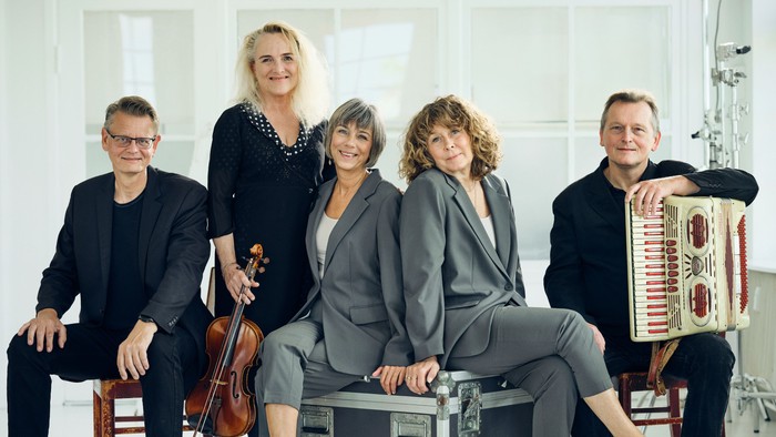 En kvintet af musikere poser sammen med instrumenter, herunder en cello, harmonika og guitarer.