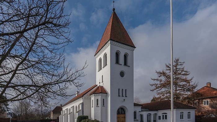 En hvid kirke med klokketårn og røde tag, omgivet af træer og en parkering med biler.