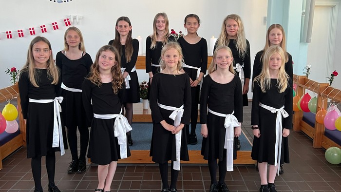 Gruppe af unge piger i formelle sorte kjoler med hvide bælter, der poserer indendørs til et foto.