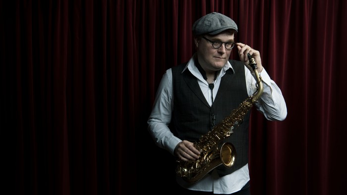 En mand i vest og kasket spiller saxofon på scenen med et eftertænksomt udtryk.