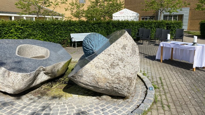 Moderne stenskulptur i en udendørs gårdhave med overdækket siddeplads og borde i nærheden.