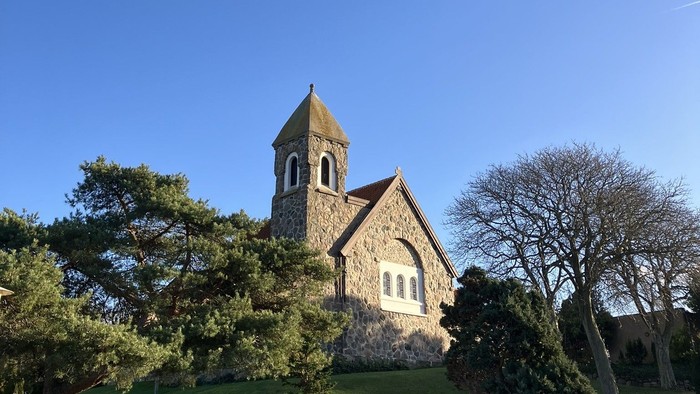 Hyggelig stenkirke med klokketårn omgivet af veltrimmet grønne områder under en klar blå himmel.