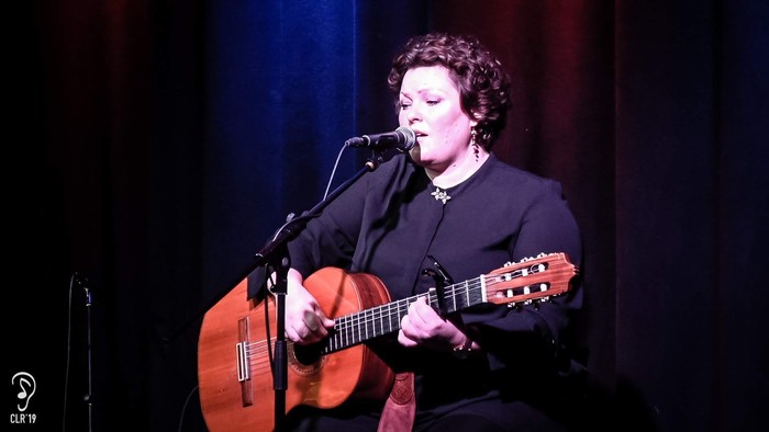 En kvinde synger lidenskabeligt i en mikrofon, mens hun spiller på en akustisk guitar på scenen.