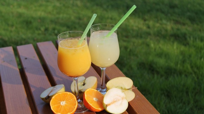 Zwei Gläser Orangensaft mit Strohhalmen und geschnittenen Orangen und Äpfeln auf einem Holztisch.
